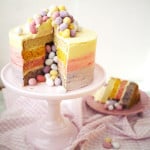 A colourful four-layer Mini Egg Cake, with piñata-style filling of mini eggs, on a cake stand with a piece on a plate, ready to eat.