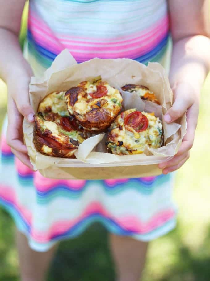 Mini Crustless Quiches with Cherry Tomatoes in a lunchbox lined with baking paper held in the hands of a little girl.
