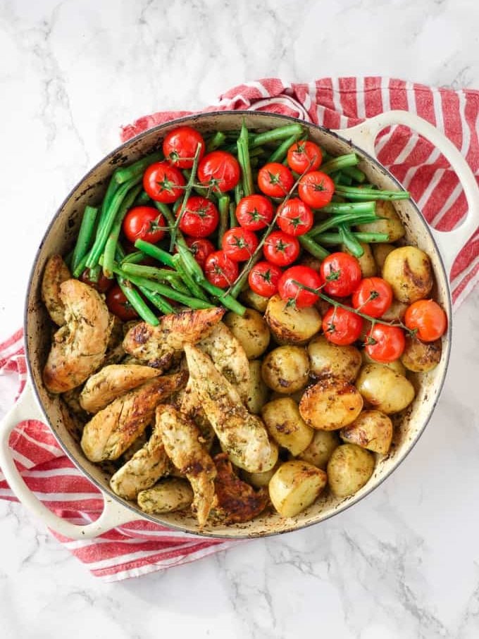 A pan on a tea towel on a white surface with baked pesto chicken and potatoes, with green beans and tomatoes.