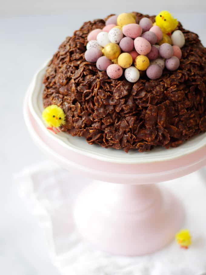 Giant Easter nest cornflake cake, circular with Mini eggs in the centre, small fake yellow chicks on a white plate and a pink stand.