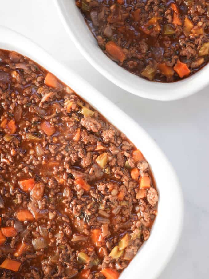 Two dishes with cottage pie base, cooked mince in, white dishes on a white background.