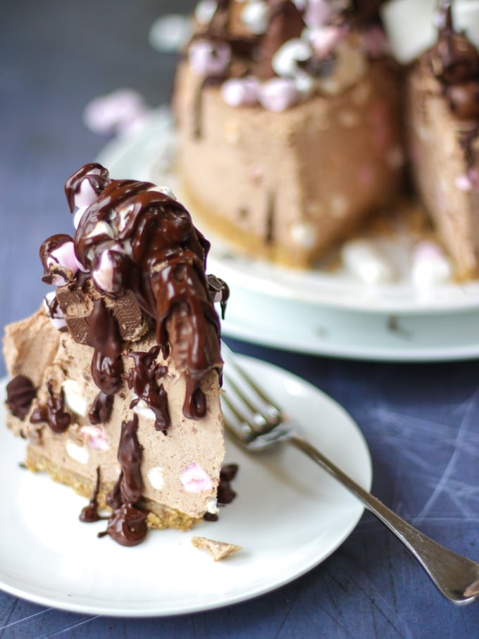 Slice of no bake rocky road cheesecake, chocolate cheesecake topped with marshmallows and chocolates and drizzled with chocolate on a white plate with a fork.