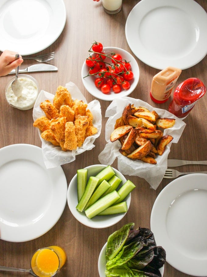 Overhead shot of homemade fish fingers, with bowls of potato wedges, tomatoes and cucumber. How to make easy peasy Homemade Fish Fingers using our simple recipe. Just four ingredients and supper tasty family meal every time.
