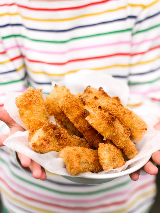 Close up of hands holding a bowl of homemade fish fingers with stripe t-shirt in the background. How to make easy peasy Homemade Fish Fingers using our simple recipe. Just four ingredients and supper tasty family meal every time.