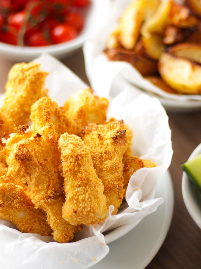 Close up of homemade fish fingers in white bowl with greaseproof paper and potato wedges in the background. How to make easy peasy Homemade Fish Fingers using our simple recipe. Just four ingredients and supper tasty family meal every time.
