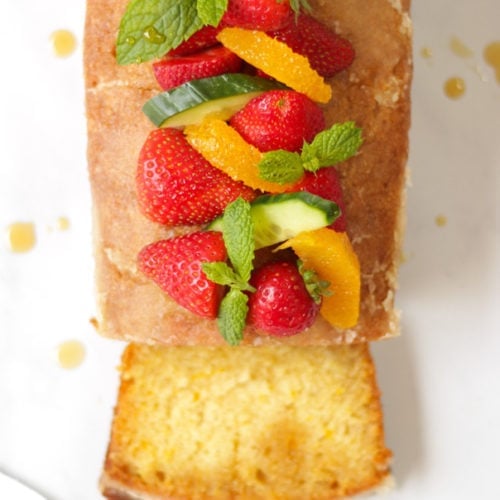 Overhead photo of Orange Drizzle Cake with Pimms. Loaf cake from overhead, topped with strawberries, mint, oranges and cucumber pieces, with slice cut off the cake.