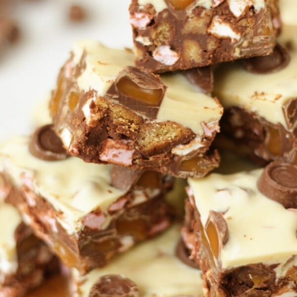 Side angle photo of a pile of Rolo Rocky Road Squares made with Roll Rocky Road Recipe, topped with white chocolate and Rolls on a wooden board.