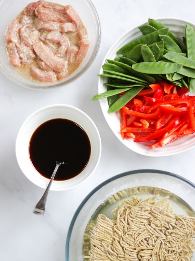 Overhead shot of pork strips, mange tout, red peppers, noodles and soy sauce and oyster sauce on white marble background for pork stir fry recipe.