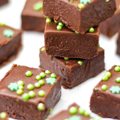 Pile of 3 pieces of easy baileys fudge and other pieces on white background with green sprinkles.
