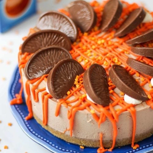 Side angle photo of a No Bake Terry's Chocolate Orange Cheesecake on a blue plate with a chocolate orange in a box in the background, drizzled with white chocolate coloured orange for this dessert recipe.