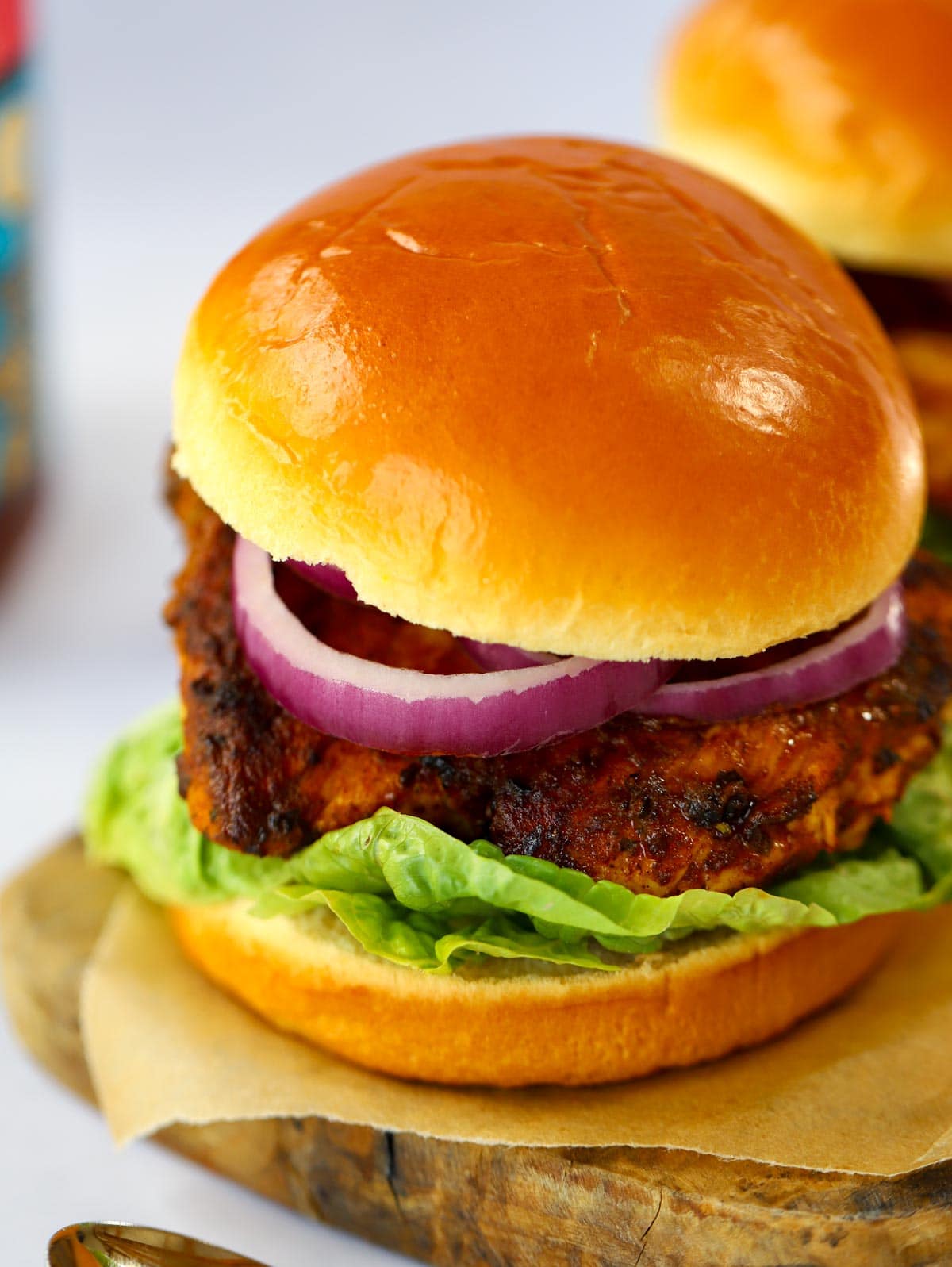 A side view of a chicken tikka burger with lettuce and red onion, served on a wooden board.