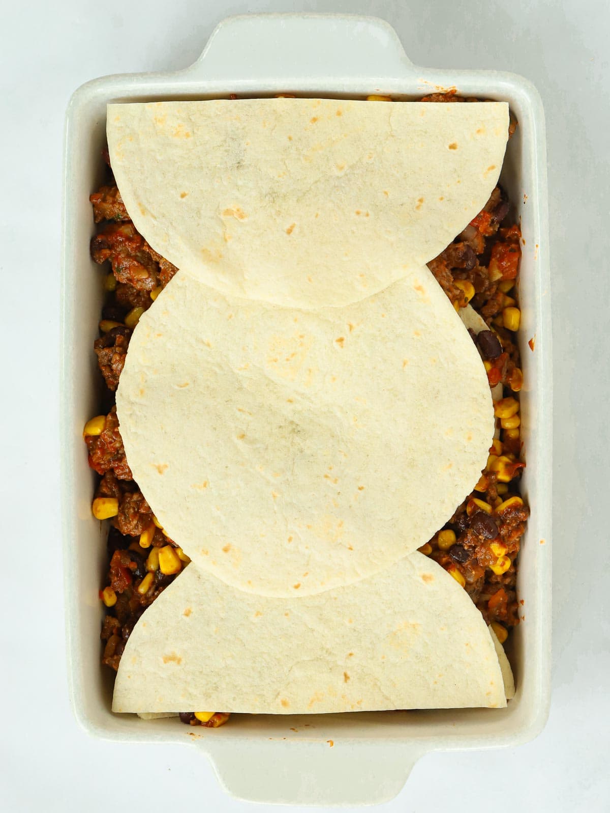 Beef mince mixture topped with uncooked tortillas in a white dish.