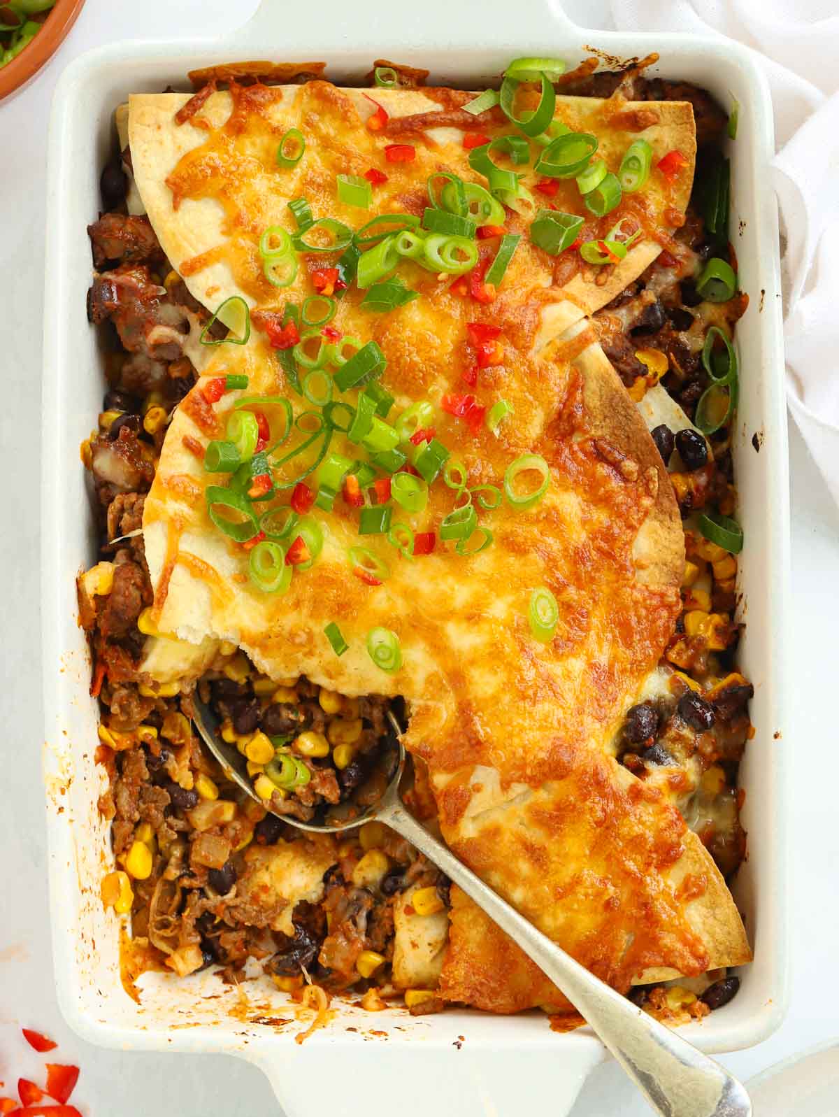 Mexican chilli beef in a white dish topped with cheese, spring onions and chilli with a spoon lying in the dish.