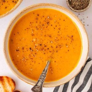 A bowl of butternut squash soup with black pepper on top and a spoon in the bowl, with toasted bread to the side, a pinch pot of black pepper.