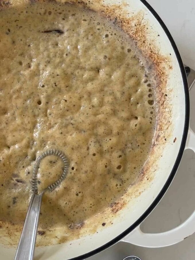 A creamy sauce cooking in a pan with a whisk in.