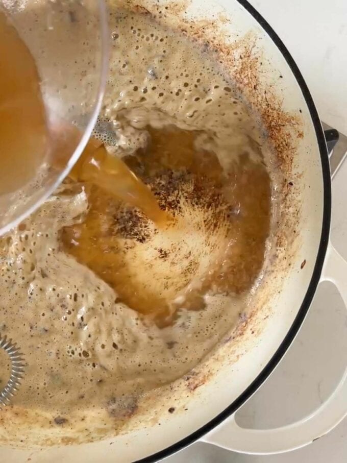 Beef stock being poured into a creamy sauce in a pan.