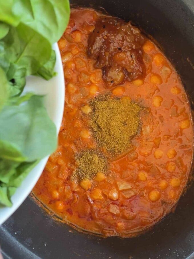 A slow cooker pan filled with chickpeas in a sauce, with spices on top and spinach about to the tipped in.