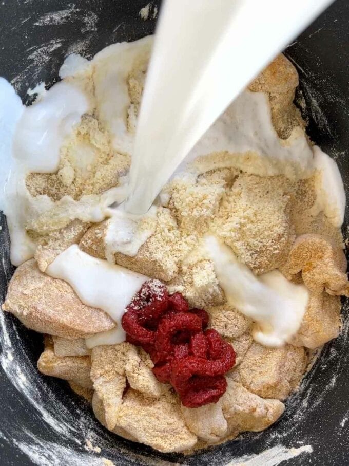 Coconut milk being poured into a slow cooker pan with chicken and tomato puree.