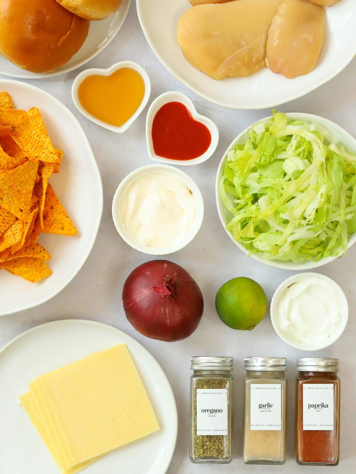 Ingredients laid out on a white surface, including sliced cheese, burger buns, lettuce, tortilla chips and spices.
