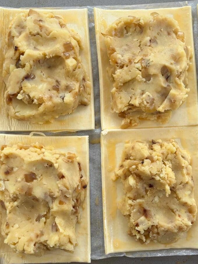 Four pastry rectangles with a cheese, potato and onion filling on top of them, with egg wash around the side of the pastry.