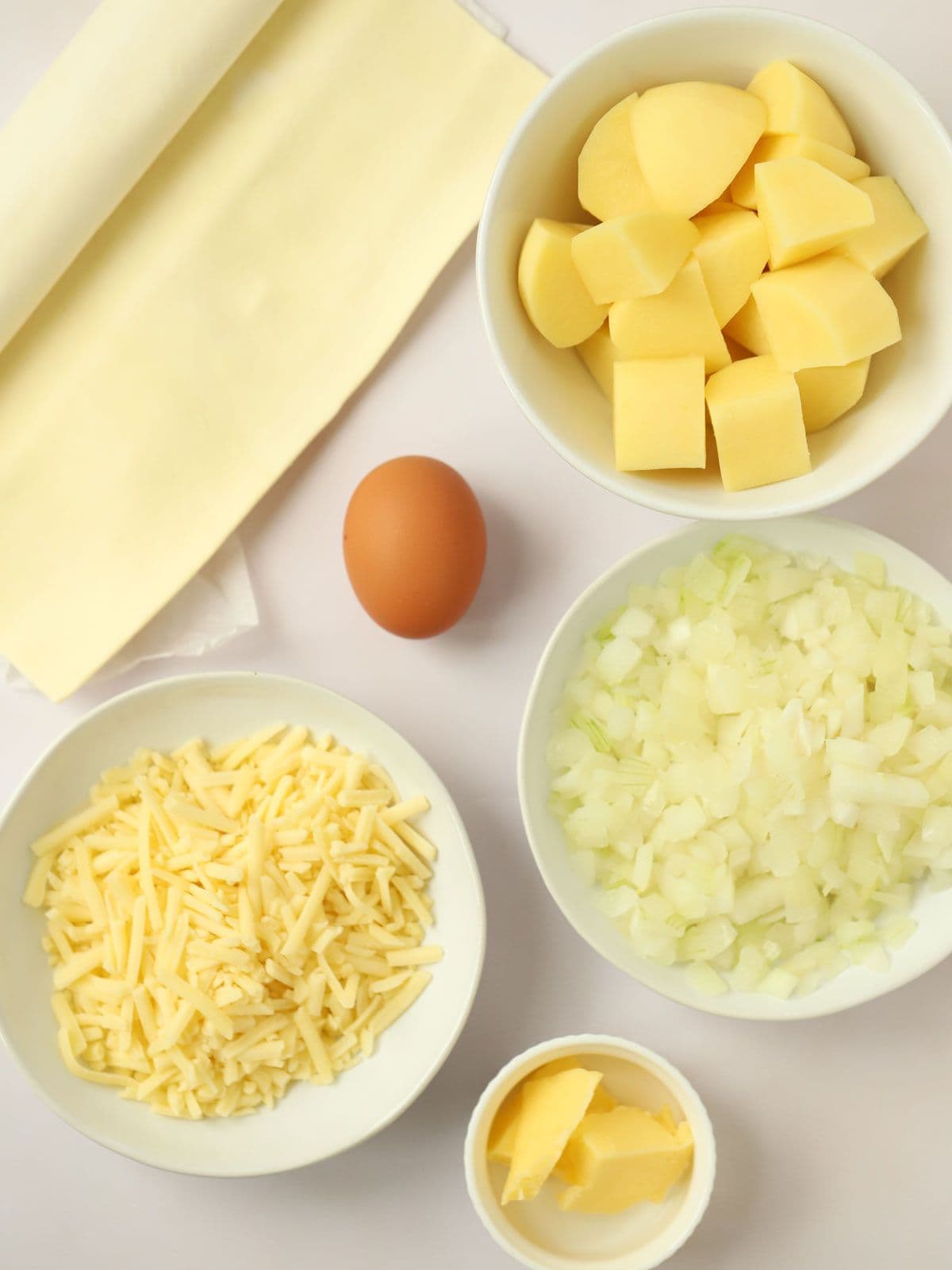 Ingredients of pastry, potatoes, egg, cheese, chopped onion and butter on a white counter top.
