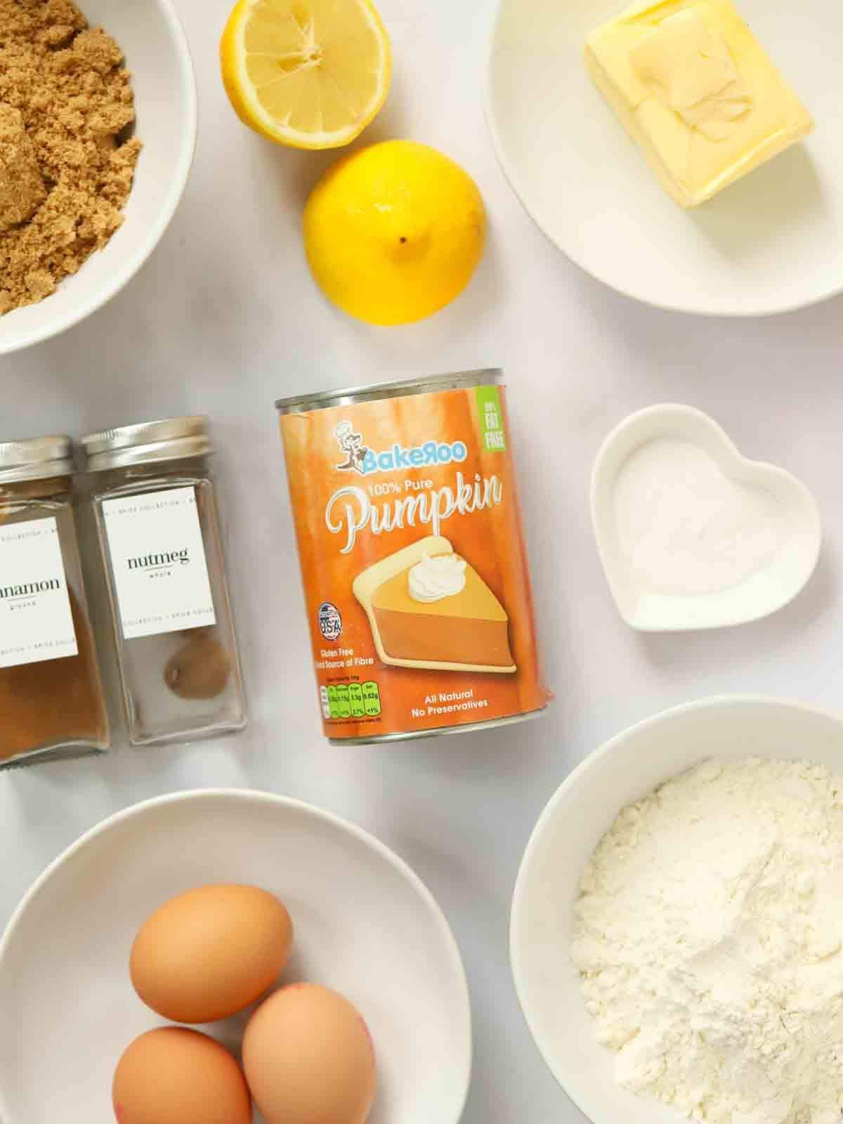The ingredients for Pumpkin Cake laid out on a counter. A tin of pumpkin puree, lemon, butter, sugar, flour, eggs and cinnamon and nutmeg spices.