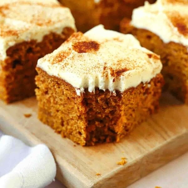 A small bit taken out of the corner of a square of pumpkin cake with cream cheese frosting on top. Other squares in the background on a board.