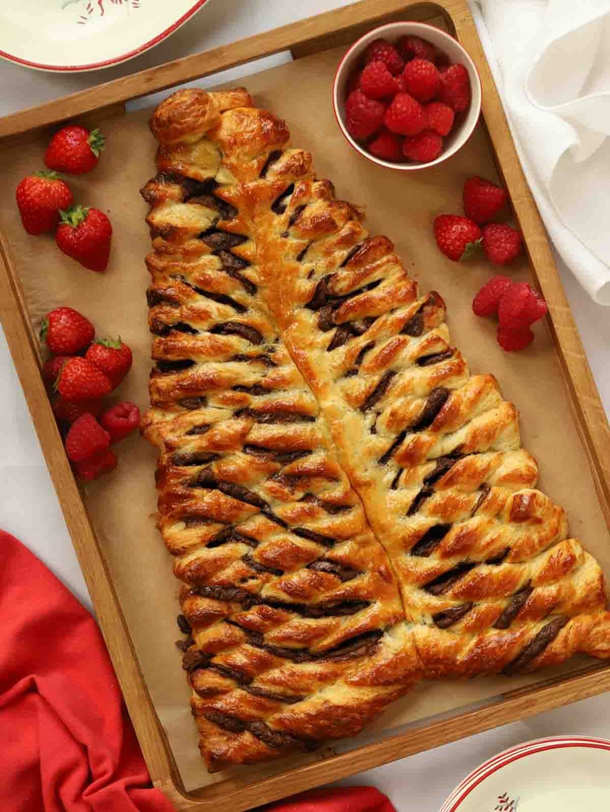 A baked Nutella Christmas Tree breakfast pastry on a wooden board, surrounding by raspberries and strawberries for a very festive breakfast.