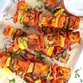 A tray with cooked paneer tikka skewers on and a bowl of raita to the side.