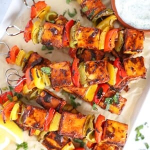 A tray with cooked paneer tikka skewers on and a bowl of raita to the side.