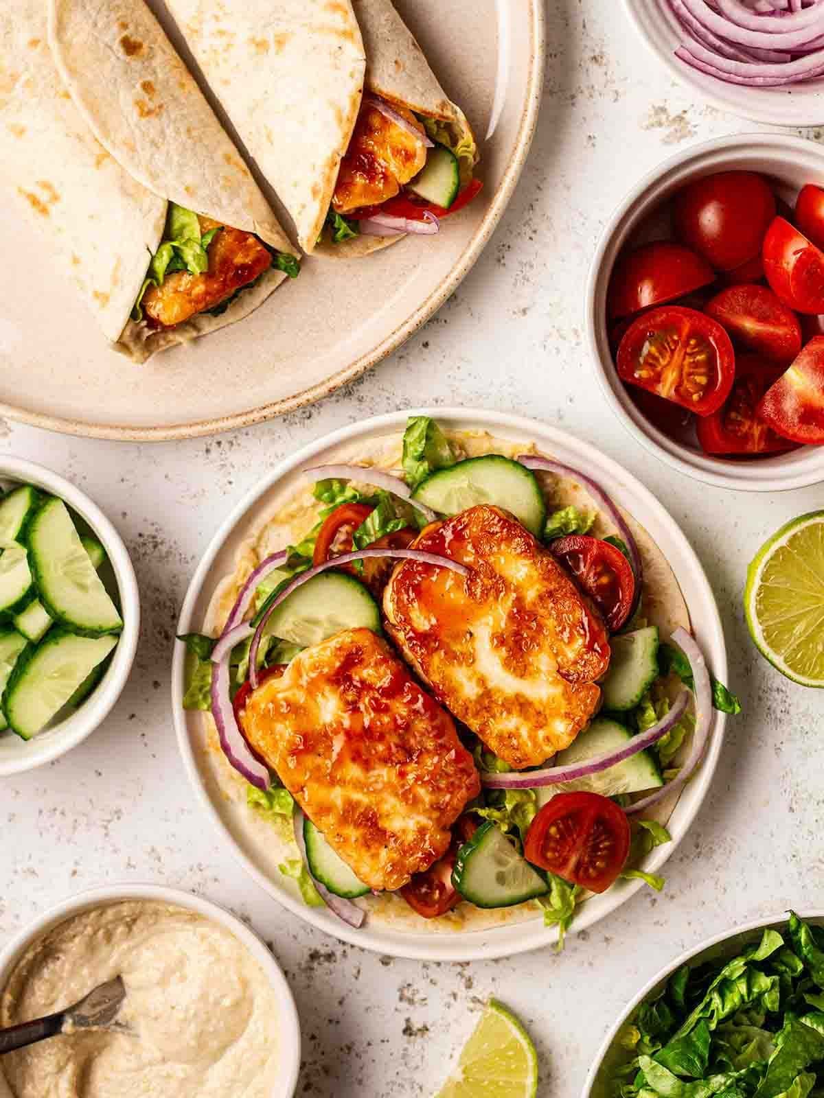 A bird's eye view of halloumi wraps with bowls of salad fillings, including tomatoes, onion and cucumber.