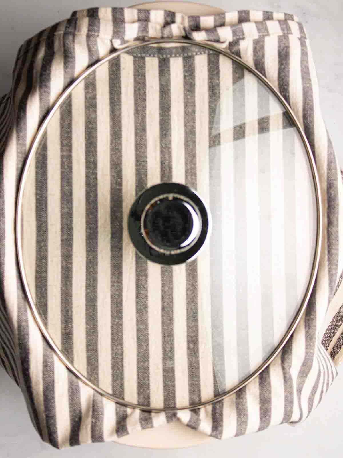 A bird's eye view of a slow cooker lid with a tea towel under it for for step 3 in the recipe for Hot Cross Bun Bread and Butter Pudding.