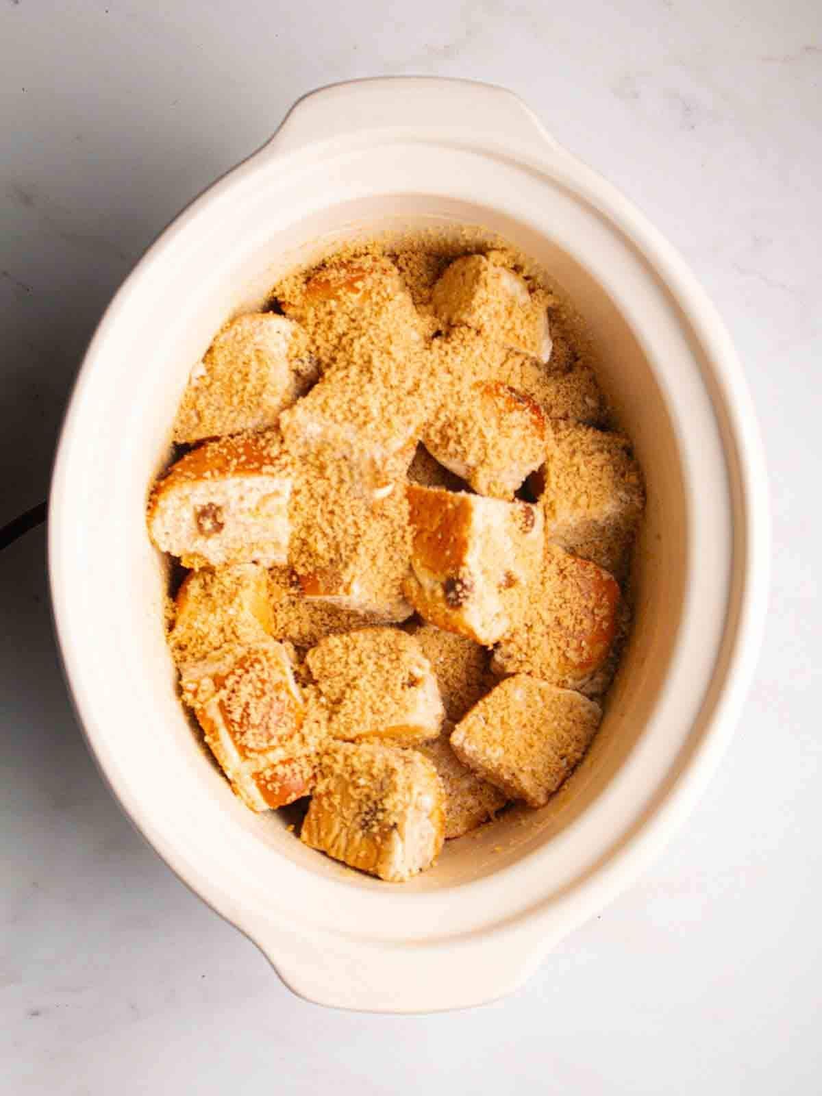 A slow cooker pan filled with pieced of chopped up hot cross buns and sugar sprinkled over the top, for step 1 in the recipe for Hot Cross Bun Bread and Butter Pudding.