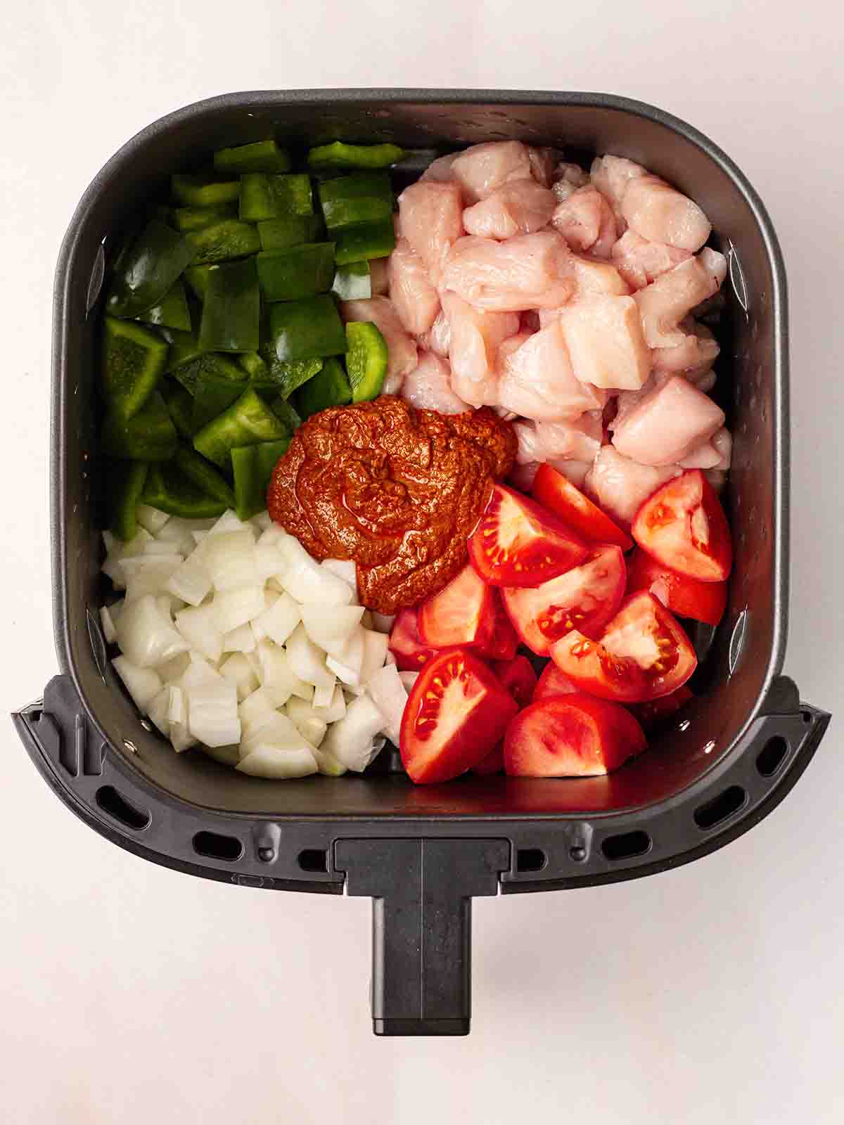 Raw ingredients in an air fryer for a chicken tikka recipe. Chicken pieces, tomatoes, green pepper, onion and tikka paste.