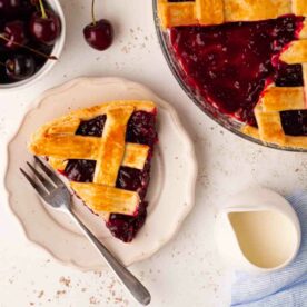 A slice of cherry pie on a plate with a fork, with the rest of the pie to the side, and a bowl of cherries, and a jug of cream on the side.