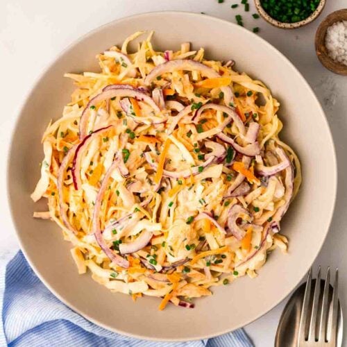 A bowl of coleslaw on a white table, with a blue cloth underneath, cutlery to the side and two small bowls above it, with chives and salt in.