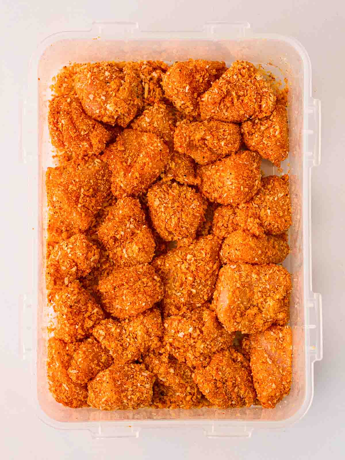 Coated pieces of raw chicken breast with breadcrumbs in a Tupperware for the recipe homemade chicken nuggets.