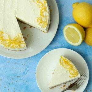 A slice of Lemon Cheesecake, with the rest of the dessert to the side, and lemons to the other side.