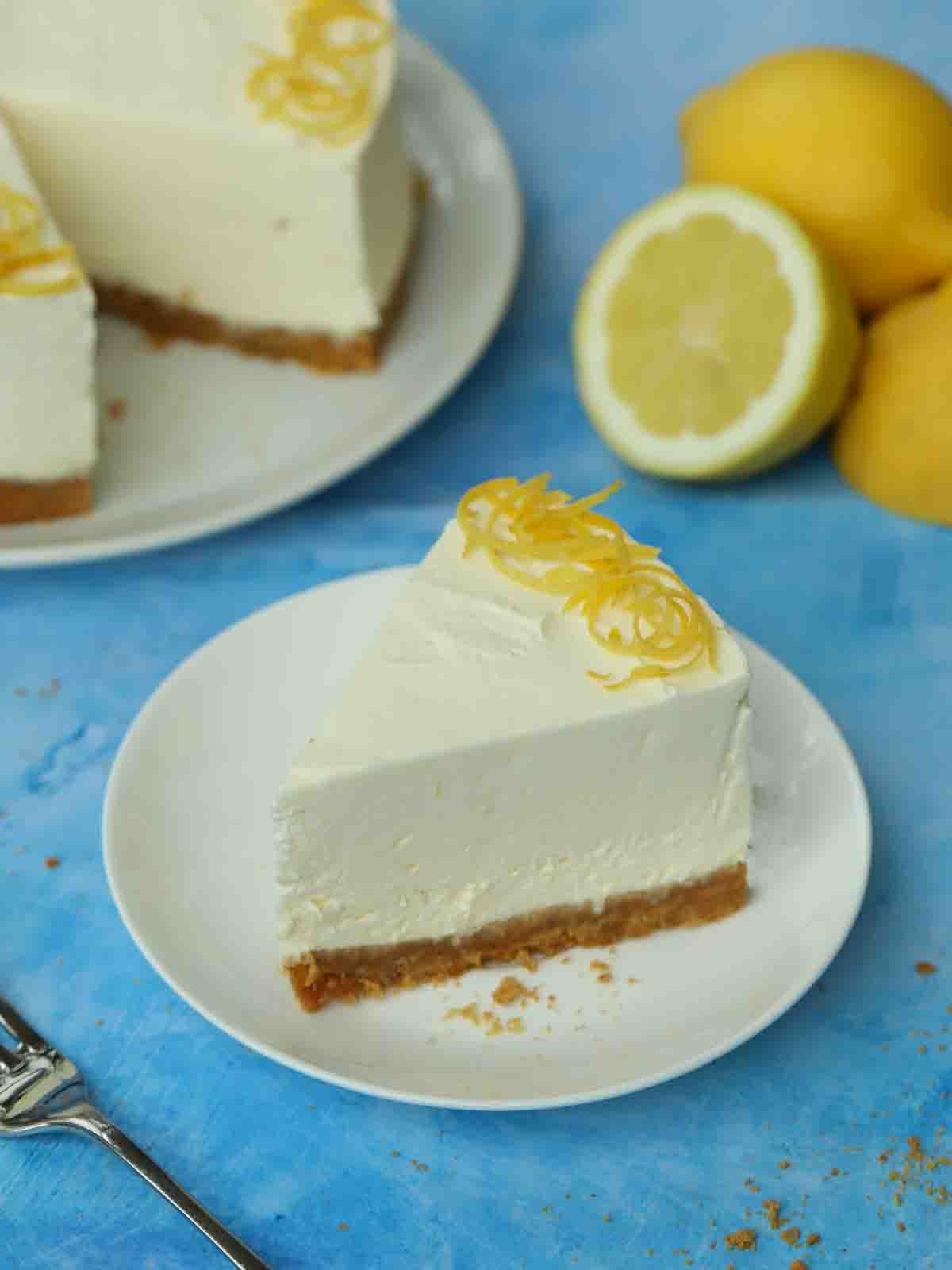 A plate with a slice of Lemon Cheesecake on, on a blue table with the rest of the cheesecake and some lemons in the background.