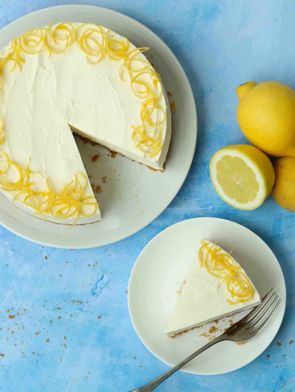 Looking down on a blue table with a lemon cheesecake, a slice taken out of it, and the slice on a plate with a fork. Lemons are in the background.