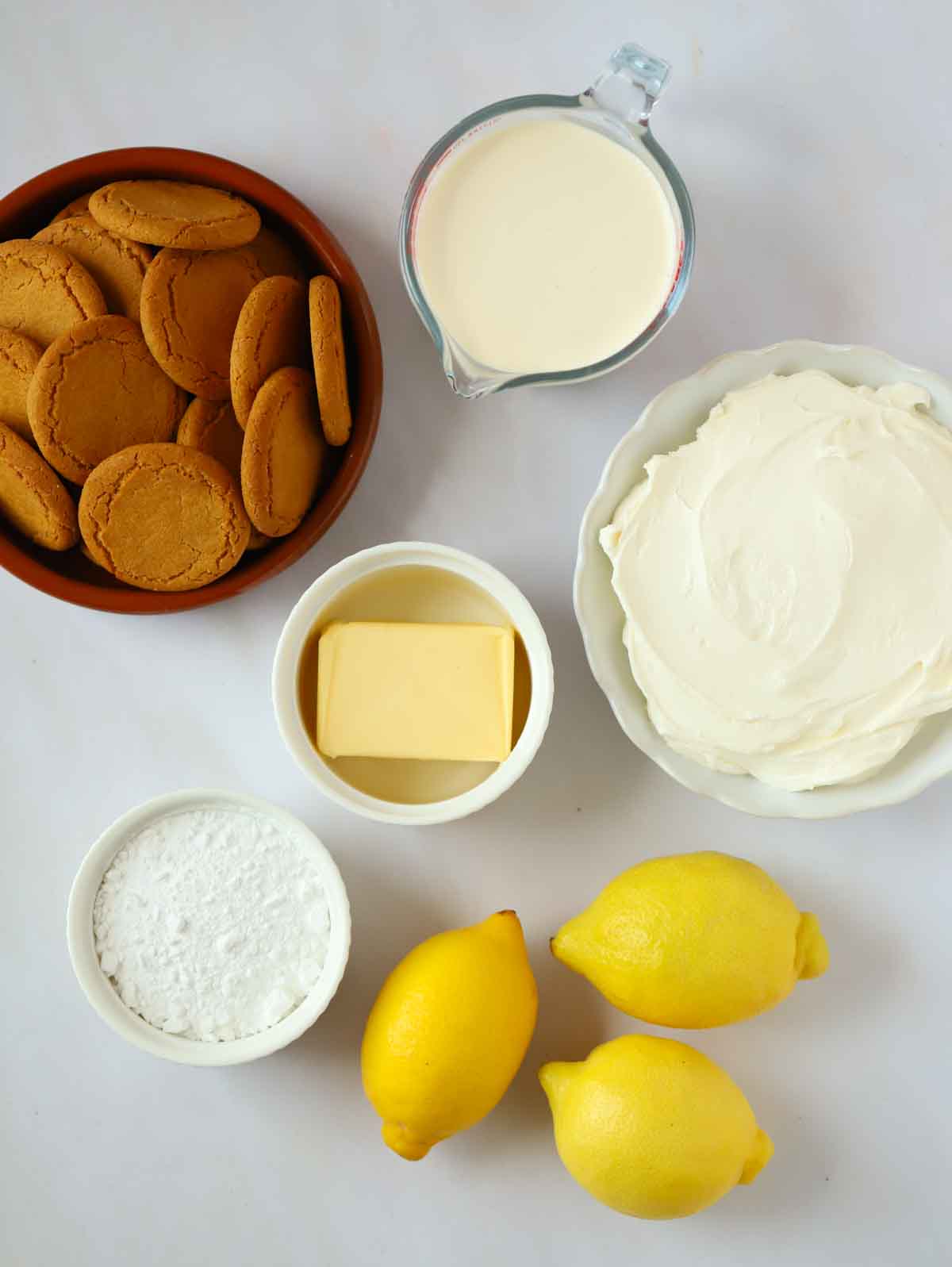 Ingredients laid out in small bowls on a white counter, including biscuits, cream and lemons.