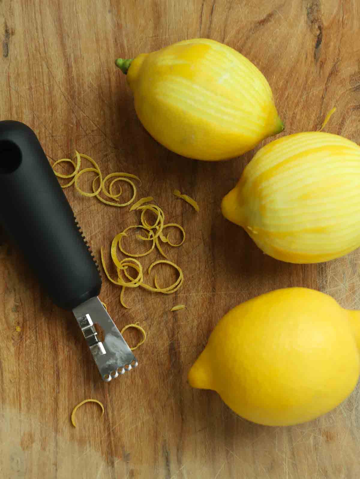 Three lemons on a wooden chopping board with a zester to the side, with some lemon zest on the board too.