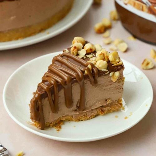 A slice of chocolate Nutella Cheesecake on a white plate with a Nutella pot in the background and the rest of the cheesecake.