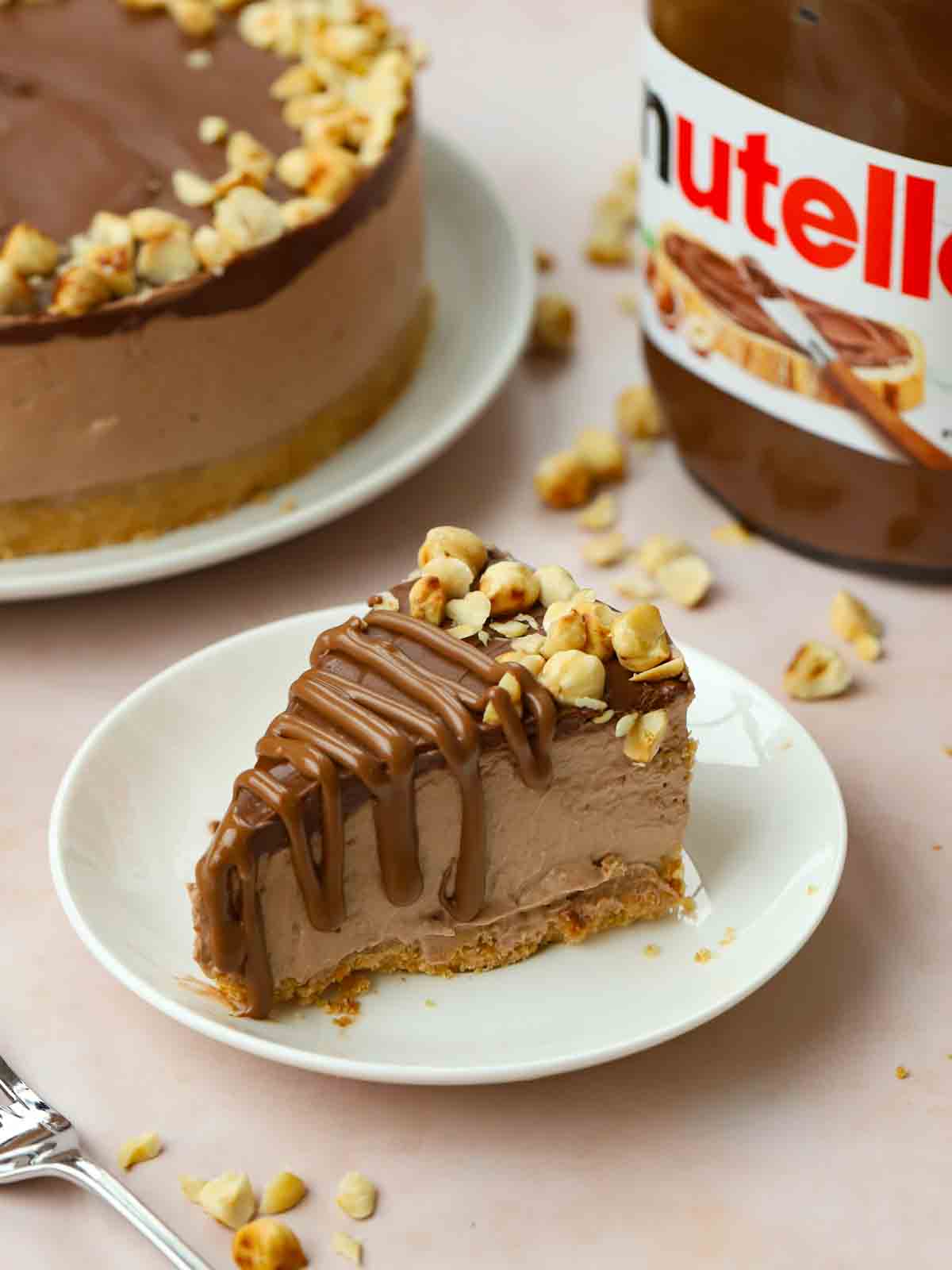 A chocolatey slice of Nutella Cheesecake on a plate, ready to eat, with the rest of the bake in the background.