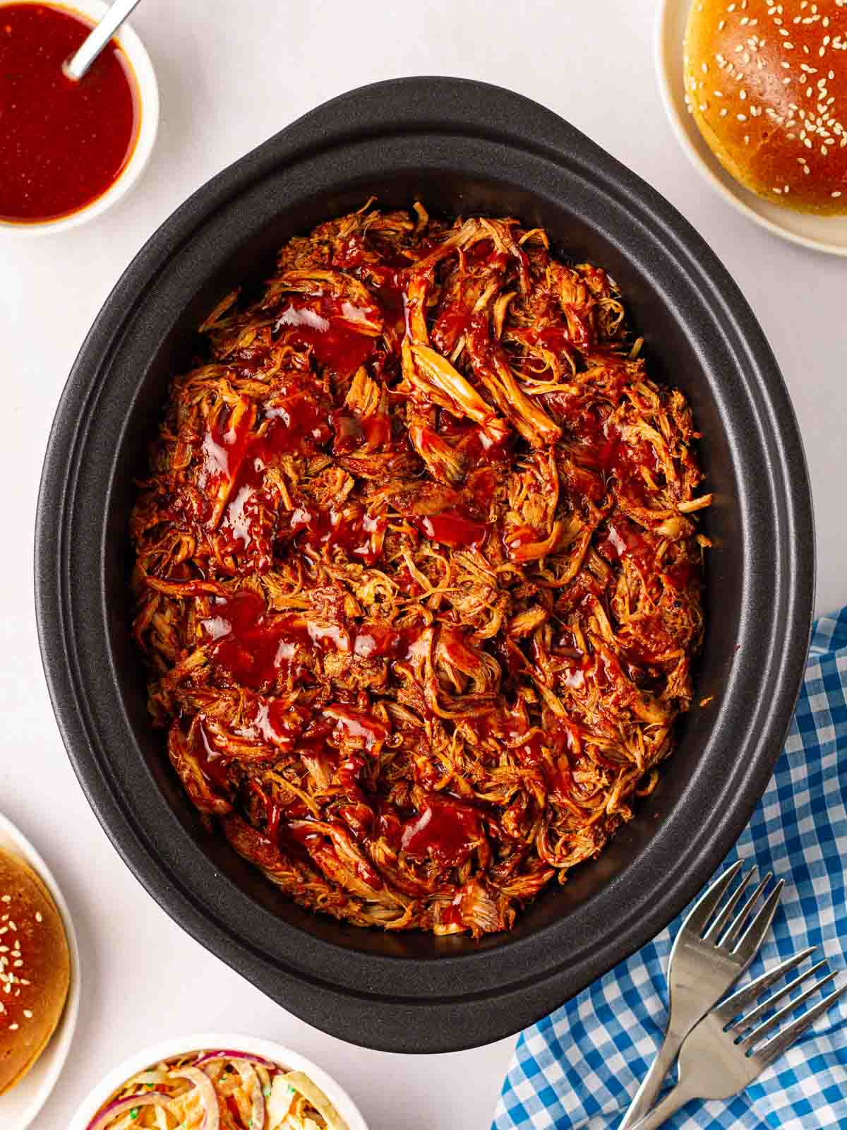 A slow cooker pan on a white tablecloth, filled with cooked pulled pork, with sauce on top and small bowls of condiments to the side.