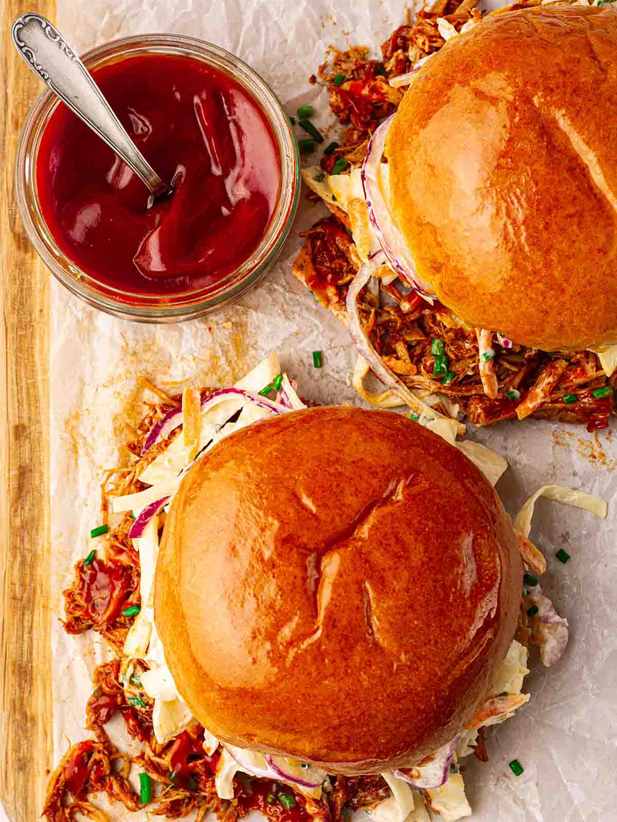 Two brioche buns filled with pulled pork and coleslaw, on a serving board with ketchup in a bowl on the side.