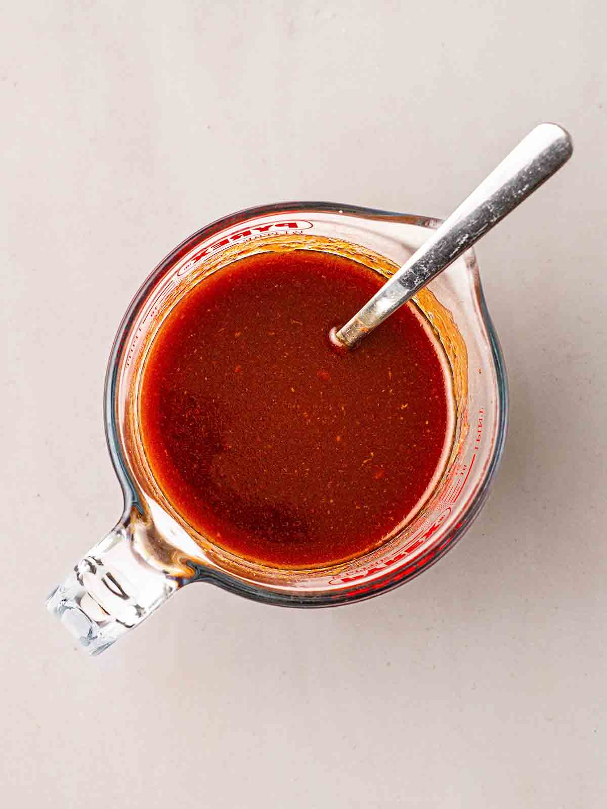 A red sauce in a glass Pyrex jug with a spoon to stir for step 2 in the process of making slow cooker pulled pork.