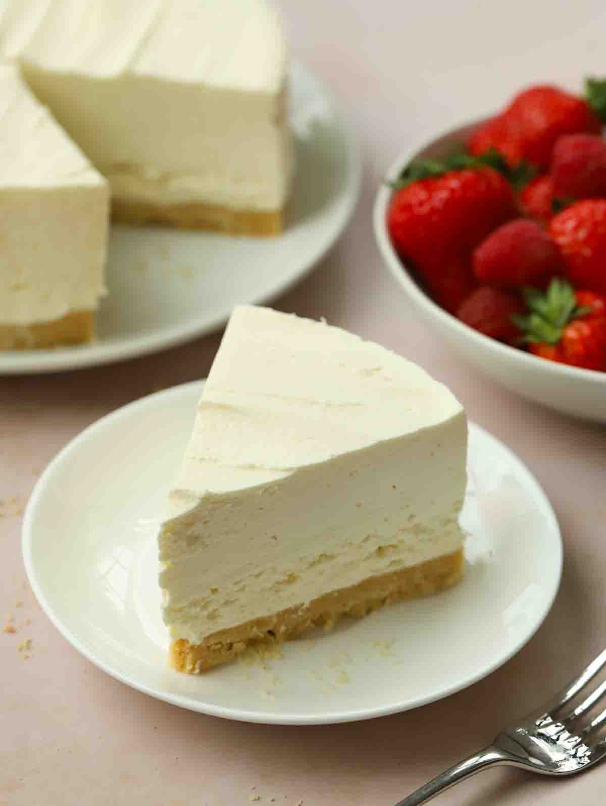 A slice of plain vanilla cheesecake on a plate, with the rest of the cheesecake and bowl of strawberries behind it.