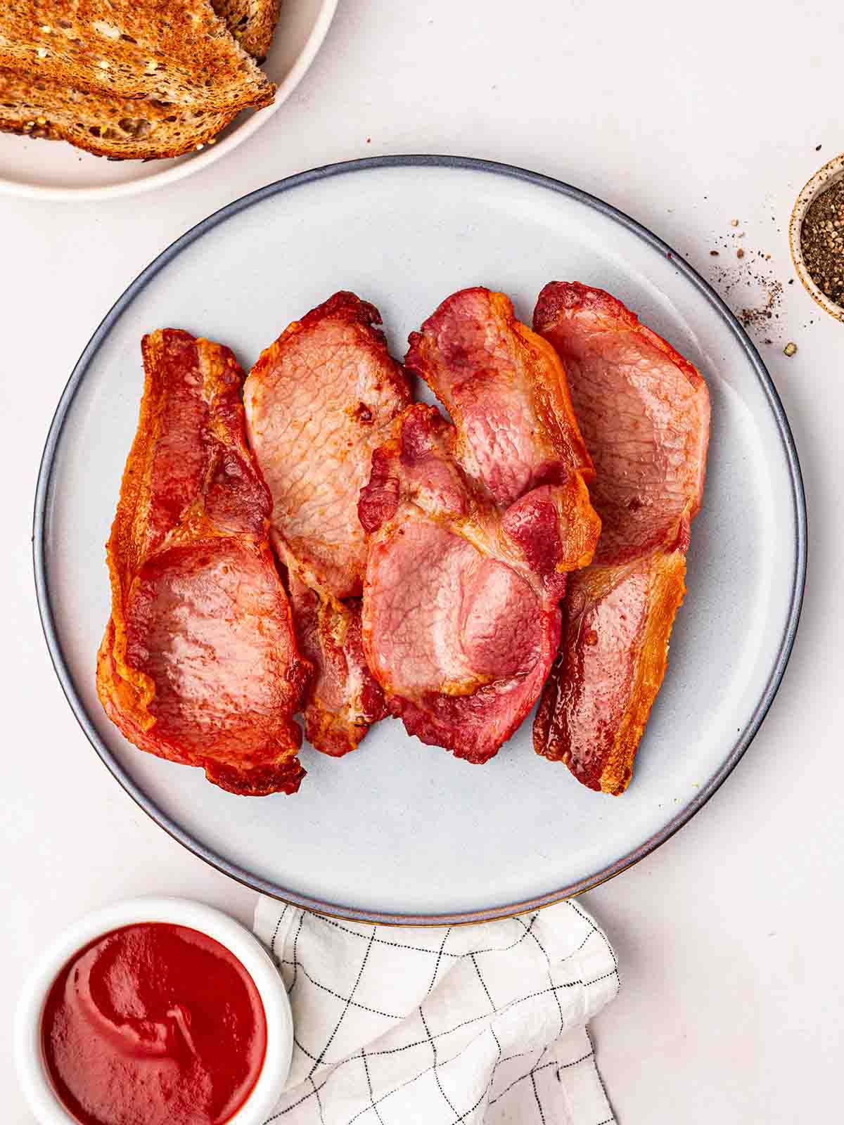 A plate with cooked bacon, on a table with toast to the side, ketchup in a bowl and black pepper.