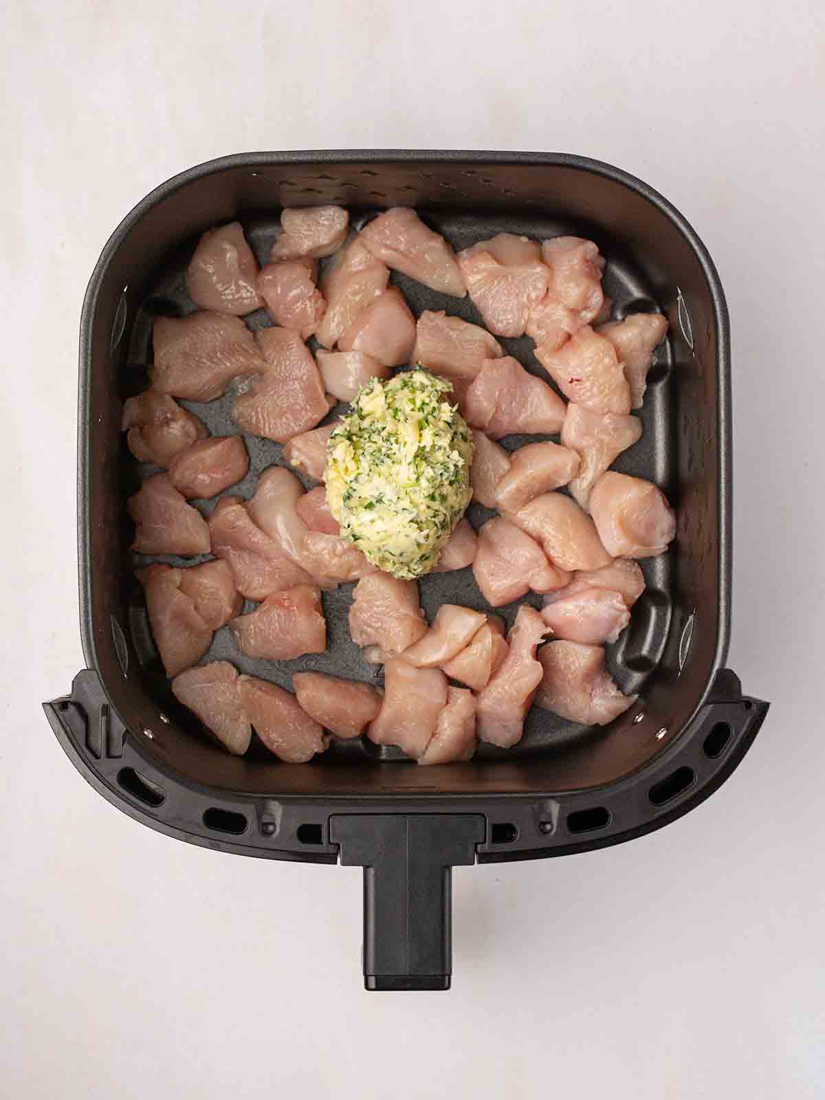 Raw chunks of chicken in an air fryer with a dollop of homemade garlic butter on top for step 1 in the recipe for Air Fryer Garlic Butter Chicken.